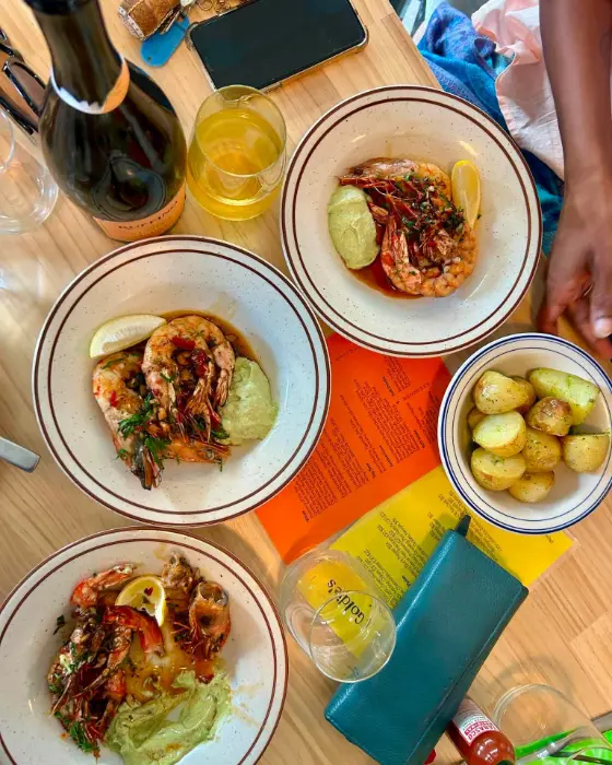 Plates of prawns and new potatoes shared across a pine table, with wine and Goldie's orange and yellow menus